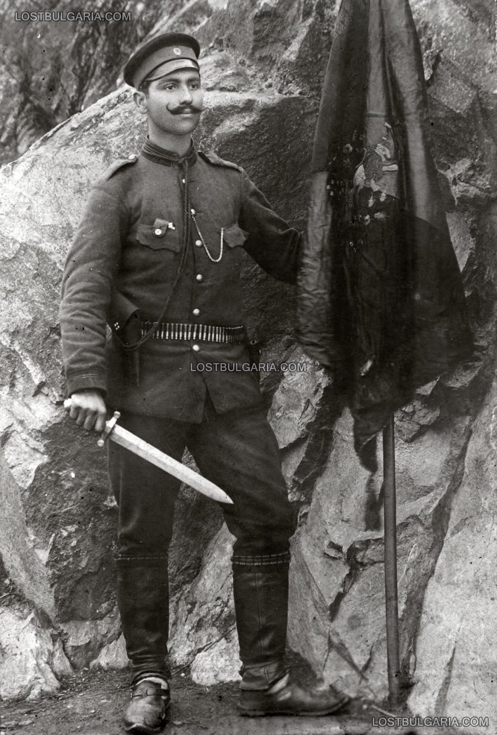 Знаменосец със знамето на 10 Прилепска дружина на Македоно-Одринското опълчение от Балканската война позира с изваден нож - тесак, датирана 14 август 1913 г.